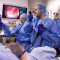 Dr. Corey Miller (centre) performs an endoscopic procedure to treat a patient who has pancreatic cancer. With his left hand (holding the black device), he guides an endoscope through the patient’s mouth and into the stomach to produce an on screen image of the the nearby tumour. With his right hand (holding the white rod), he delivers a sliver of metal—known as a “source”, which emits alpha particles—into the tumour. Assisting Dr. Miller are Dr. Nicolas Chapelle (left), an Advanced Endoscopy Fellow, and Brandon Ignacz, an Advanced Endoscopy Nurse. (Click on this photo to enlarge it.)